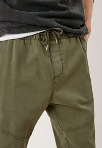 Man in olijfgroene broek met trekkoord en een zwart shirt met een wit hemd eronder, hand in de zak.