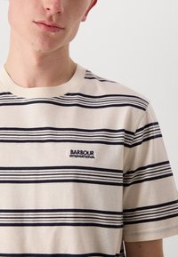 Kreemikas T-särgid siniste horisontaalsete triipudega, ümmargune kaelus ja tikitud "Barbour International" logo rinnal.