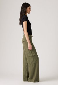 Donna in una maglietta nera attillata a maniche corte e pantaloni cargo verdi oliva a gamba larga, in profilo su uno sfondo semplice.