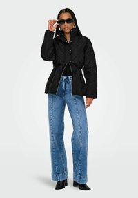 Veste matelassée noire avec une taille cintrée, dotée d'un col haut et de quincaillerie en argent, associée à un jean bleu clair taille haute et des bottes noires.