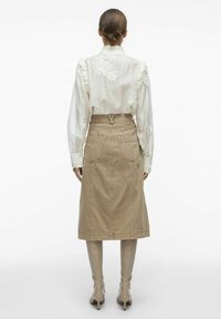 Blusa blanca con bordado floral, combinada con una falda de mezclilla beige. La falda tiene una cintura con cinturón y bolsillos delanteros. Botas ajustadas de piel lisas hasta la rodilla.