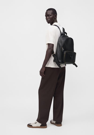 Homme portant une chemise blanche à manches courtes, un pantalon large foncé, des chaussures blanches et un sac à dos en cuir noir, debout devant un fond clair uni.