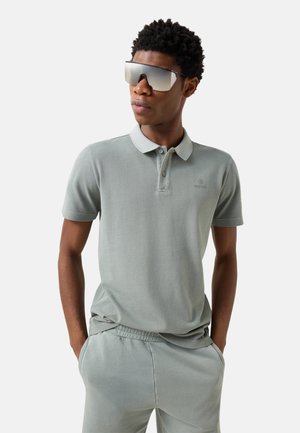 Młody mężczyzna w jasnoszarej koszulce polo, dopasowanych spodniach i dużych, refleksyjnych okularach przeciwsłonecznych, stojący z rękami w kieszeniach.