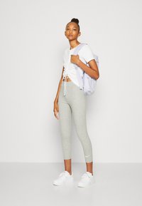 Leggings grises, camiseta corta blanca con un nudo y zapatillas blancas. Mochila morada clara. Texturas suaves, diseño ajustado.