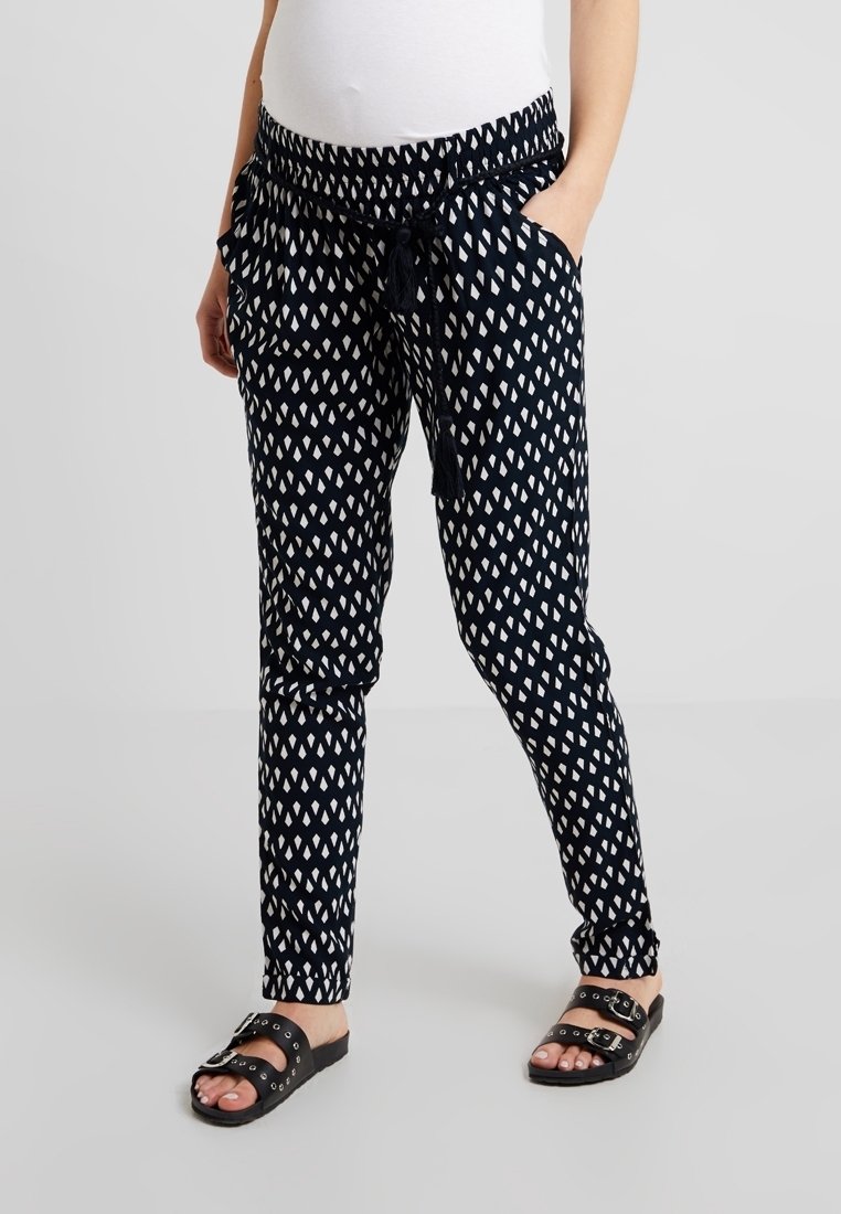 Femme portant un pantalon fuselé à motifs noir et blanc avec ceinture à pompons et sandales noires à boucles, mains dans les poches.