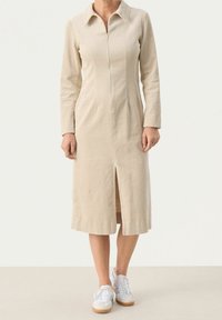 Robe en velours côtelé beige avec col, fermeture éclair à l'avant, manches longues et fente centrale à l'avant, portée avec des baskets blanches.