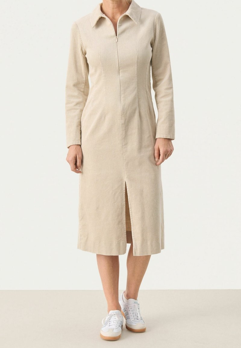 Robe en velours côtelé beige avec col, fermeture éclair à l'avant, manches longues et fente centrale à l'avant, portée avec des baskets blanches.