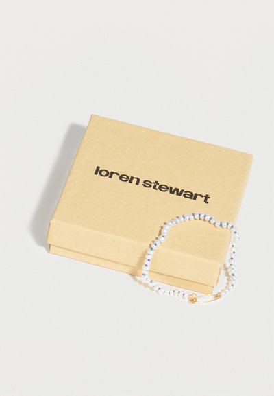 Βραχιόλι με χάντρες σε λευκό και μπλε χρώμα, με κούμπωμα πινέζας ασφαλείας σε χρυσό, τοποθετημένο σε μπεζ κουτί Loren Stewart.