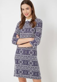 Mujer con cabello castaño sonriendo, vistiendo un vestido azul marino con un estampado blanco y un cuello blanco, con los brazos cruzados.