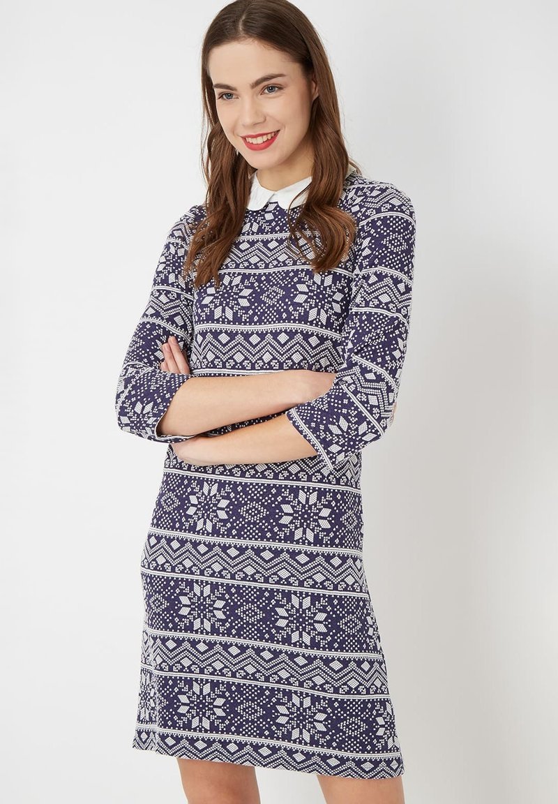 Mujer con cabello castaño sonriendo, vistiendo un vestido azul marino con un estampado blanco y un cuello blanco, con los brazos cruzados.