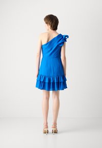 Robe bleue à une épaule en tissu fluide, avec un accent volant à l'épaule et un ourlet à volants superposés. Les sandales sont beiges.