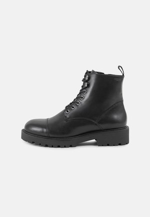 Botas de cuero negras hasta el tobillo con puntera redondeada, parte delantera con cordones, suela de goma gruesa y una lengüeta en la parte posterior para facilitar su uso.
