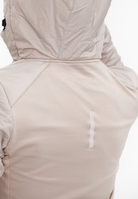 Lysbeige hættejakke med en glat tekstur. Har en dekorativ vertikal pynt på ryggen og detaljer på skuldersømmen.