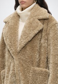 Femme portant un manteau teddy beige duveteux par-dessus un pull col roulé blanc avec des boucles d'oreilles créoles dorées, visible des épaules jusqu'au milieu du torse.