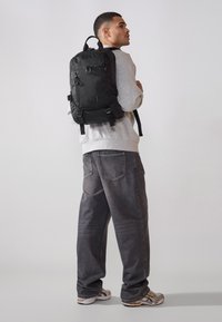 Sac à dos en tissu noir avec des sangles réglables et plusieurs compartiments, porté par une personne portant un sweat-shirt gris et un jean sombre.