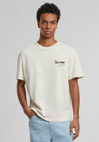 T-shirt en coton beige clair à manches courtes et col rond, avec un graphique "Votre Favori de Tous les Temps" sur la poitrine gauche.