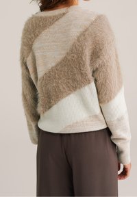 Pull beige et crème avec une texture duveteuse en motif diagonal, doté de poignets et d'un ourlet côtelés. Associé à un pantalon marron.