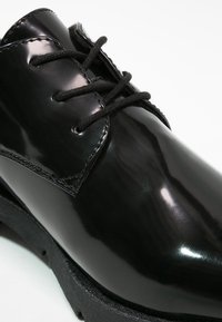 Chaussure habillée en cuir noir verni avec lacets noirs et semelle épaisse, présentée en vue rapprochée et en angle sur fond blanc.