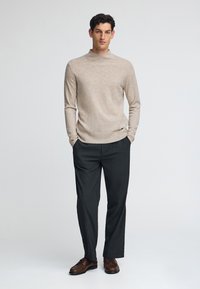 Hombre de pie con las manos en los bolsillos, vistiendo un suéter beige, pantalones gris oscuro y zapatos de cuero marrón, contra un fondo liso.