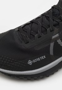 Zwarte hardloopschoen met een textuur bovenwerk, vetersysteem en GORE-TEX branding, met een grijze zool en geometrische patronen.