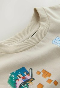 Bež pulover z okroglim ovratnikom z motivom videoigričnega lika v pikslih, ki drži meč, in majhnimi oranžnimi pikselnimi bloki na tkanini.