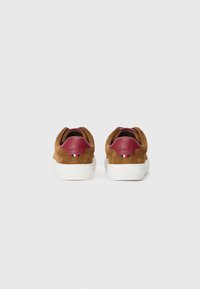 Zapatillas de ante marrón con una pestaña de talón de cuero burdeos, suela de goma blanca y un pequeño acento tricolor en la parte trasera. Textura suave.
