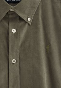 Camicia in corduroy verde oliva con colletto button-down, caratterizzata da bottoni bianchi e un logo con figura ricamata sul petto. Tessuto testurizzato.