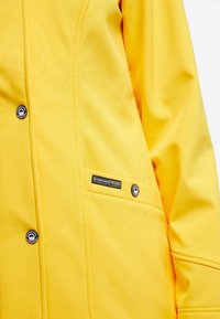Yellow waterproof jacket with a smooth texture, featuring a side pocket, circular button closure, and a branded label stitched on the chest.