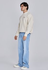 Beige sweatshirt met geborduurd logo, gecombineerd met lichtblauwe jeans. De outfit heeft een relaxte pasvorm en casual sneakers met grijze accenten.