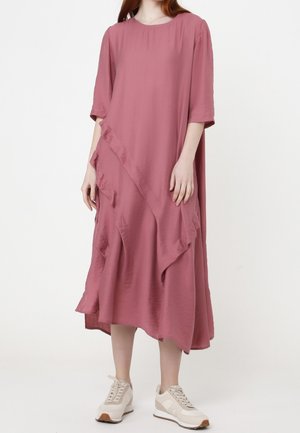 Vrouw draagt een losse, mid-lange jurk in stoffig roze met ruches en halflange mouwen, gecombineerd met beige sneakers, staand tegen een witte achtergrond.