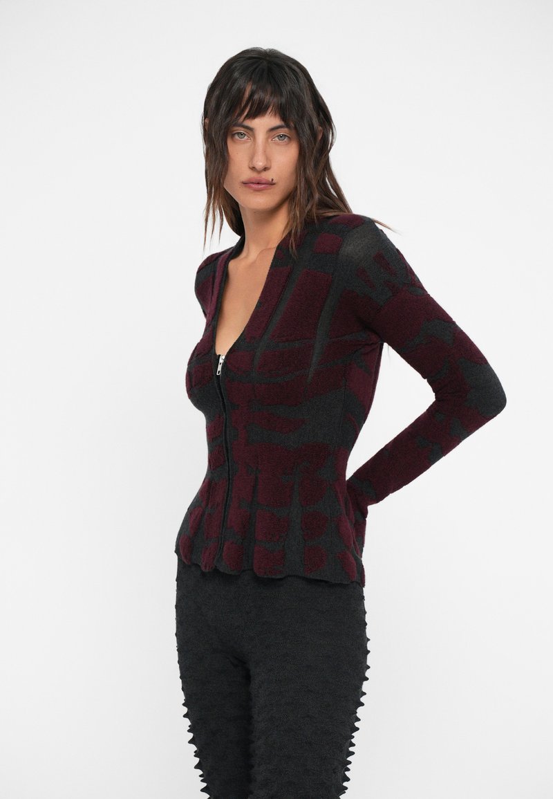Gilet en tricot gris foncé et bordeaux avec fermeture éclair à l'avant, présentant un motif abstrait et une silhouette ajustée avec une texture côtelée.