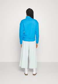 Hellblaues Sweatshirt mit hohem Kragen, oversized Passform und geripptem Saum. Kombiniert mit mintgrünen Weitbein-Hosen und weißen Turnschuhen.