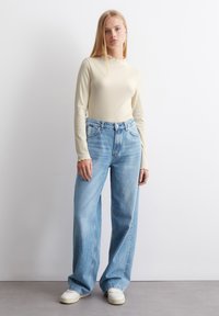 Langärmliges, geripptes Oberteil in hellbeige mit gewelltem Ausschnitt, kombiniert mit weit geschnittenen blauen Denim-Jeans und weißen Sneakern.