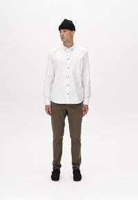 Biela košeľa s gombíkmi a vreckom na hrudi, spárovaná s hnedými nohavicami. Model má na hlave čiernu pletenú čiapku a na nohách čierne topánky. Minimalistický dizajn.