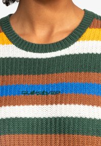 Acercamiento de un suéter de punto a rayas en verde, marrón, blanco, azul y amarillo, con cuello de canalé y nombre de la marca bordado en el pecho.