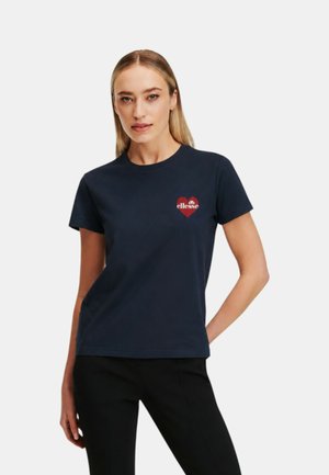Donna con capelli biondi lisci indossa una t-shirt blu navy con un cuore rosso e il logo "ellesse" sul petto sinistro, abbinata a pantaloni neri.