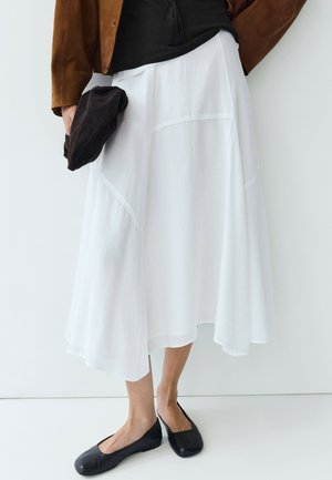 Person, die einen weißen Midi-Rock, schwarze Ballerinas, ein schwarzes Top und eine braune Jacke trägt und einen schwarzen zusammengefalteten Hut vor einem einfarbigen weißen Hintergrund hält.