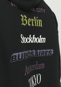 Sudadera negra con capucha que presenta nombres de ciudades en varios tipos de letra y colores: verde, púrpura y blanco sobre una tela texturizada. Diseño con capucha.