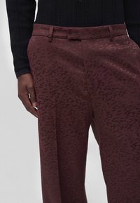 Gros plan d'une personne portant un pantalon bordeaux avec un motif léopard subtil et un haut en maille noir à manches longues.