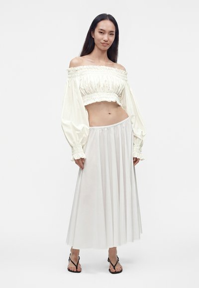 Γυναίκα στέκεται φορώντας κρεμ off-shoulder crop top με μακριά μανίκια, αέρινη λευκή φούστα και μαύρα σανδάλια, κοιτάζοντας μπροστά.