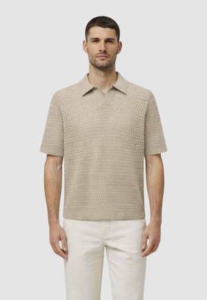 Uomo che indossa una polo beige a maniche corte in maglia testurizzata e pantaloni chiari, in piedi contro uno sfondo chiaro uniforme.