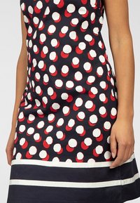 Femme portant une robe sans manches bleu marine à pois blancs et rouges, avec un ourlet bleu marine à rayures blanches, main posée sur le côté.