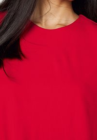 Czerwony top z tkaniny o gładkiej strukturze i szerokim, zaokrąglonym dekoltem, z minimalnymi detalami i dopasowanym fasonem.