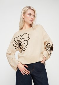 Béžový pletený svetr s velkou černou květinovou výšivkou na rukávech, s žebrovaným lemem a volným střihem. Kombinován s tmavými džíny.