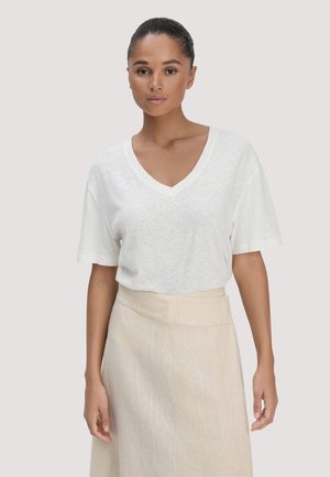 Woman with tied-back hair wearing a white V-neck short-sleeve top and a beige high-waisted skirt, standing against a plain background.