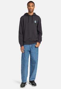 Felpa nera con tasca a marsupio frontale e cappuccio con cordini, abbinata a jeans in denim blu chiaro e scarpe sportive nere.