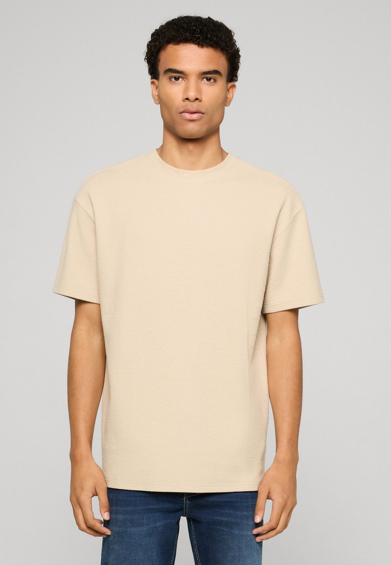 Jeune homme aux cheveux courts et bouclés portant un T-shirt beige uni à manches courtes et texturé ainsi qu'un jean bleu, debout devant un fond neutre.