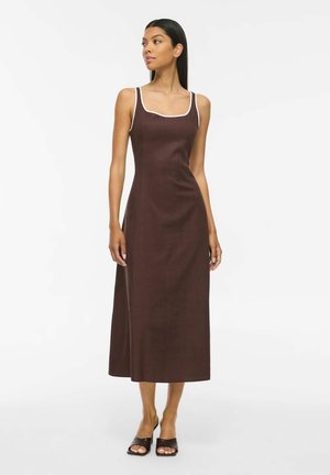 Femme portant une robe midi marron sans manches avec une bordure blanche et des sandales à talons noires à bout ouvert, debout sur fond blanc.