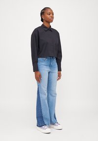 Lyhyt musta kauluspaidassa, yhdistettynä vaaleansinisiin leveälahkeisiin farkkuihin, joissa on tummempi denim-paneeli, sekä valkoisiin tennareihin.