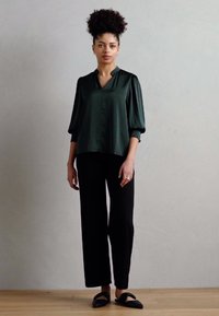 Blouse en satin vert foncé avec un décolleté en V et des manches bouffantes, associée à un pantalon noir à jambes larges et des chaussures noires à enfiler.
